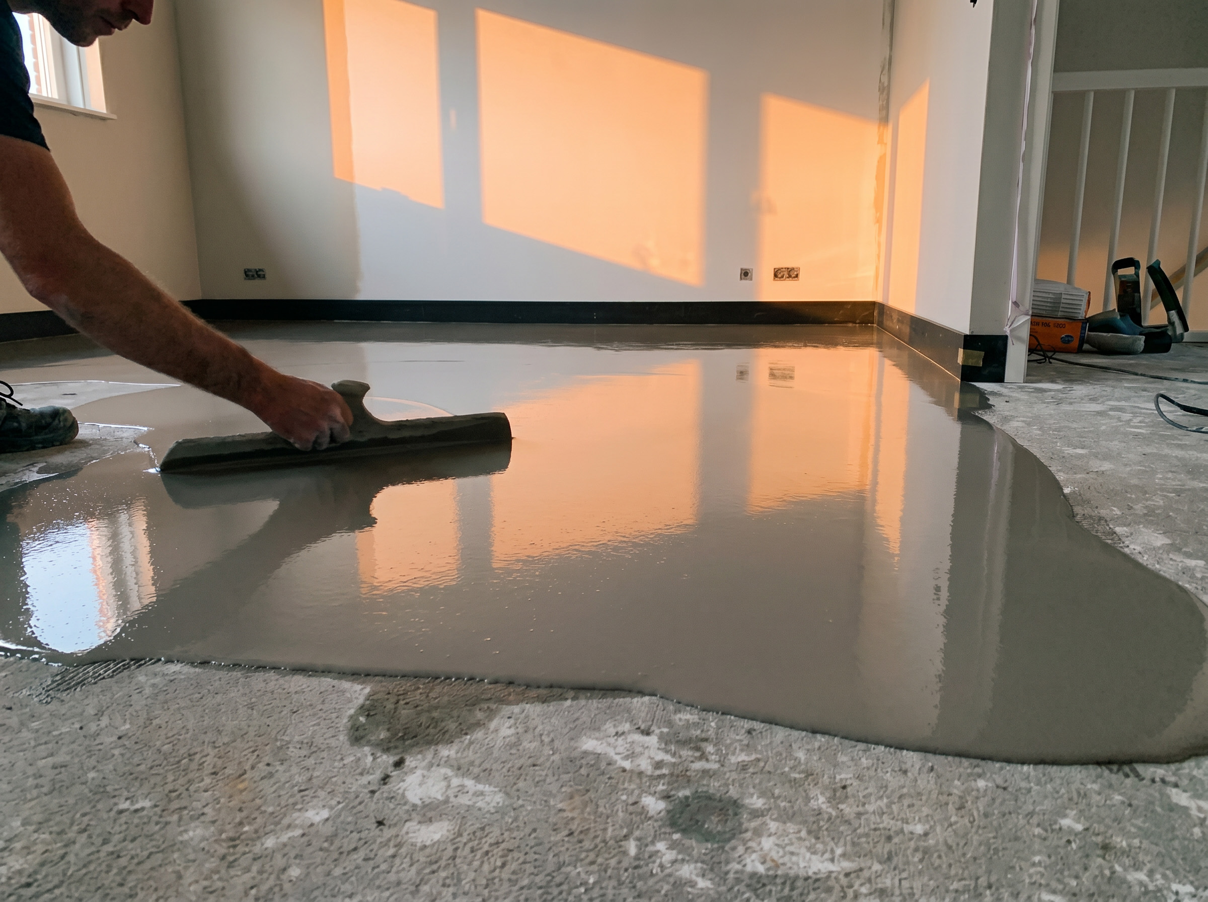 Vakman strijkt een vloeibare egalisatielaag uit over een betonnen vloer in een kamer met warm invallend zonlicht.