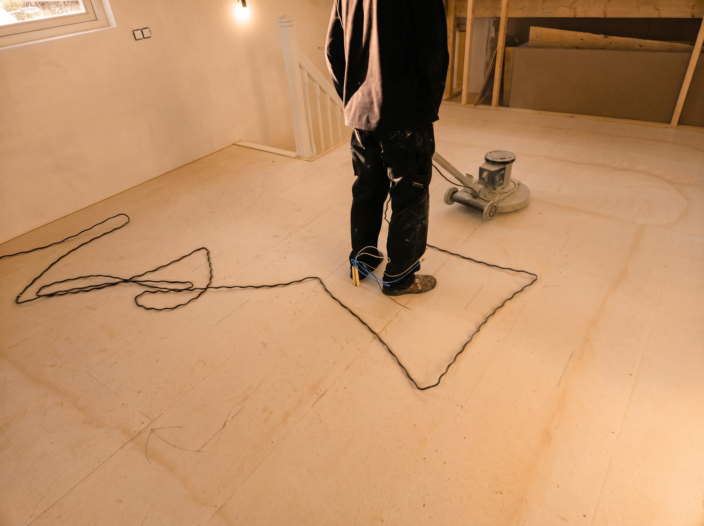 Vakman strijkt een vloeibare egalisatielaag uit over een betonnen vloer in een kamer met warm invallend zonlicht.
