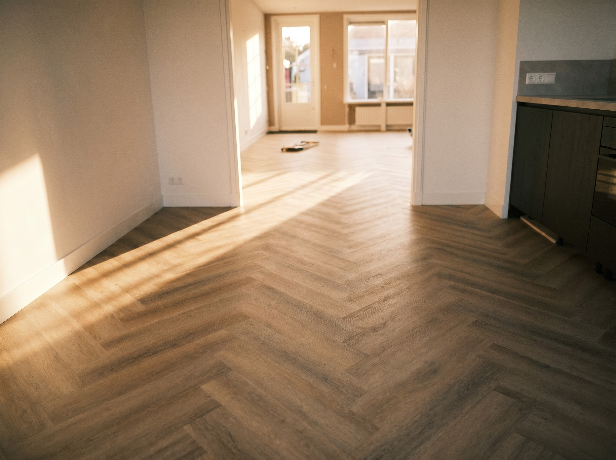 Ruime woonkamer met een strak gelegde visgraatvloer, waar warm zonlicht door de ramen naar binnen valt en de vloer mooi uitlicht.